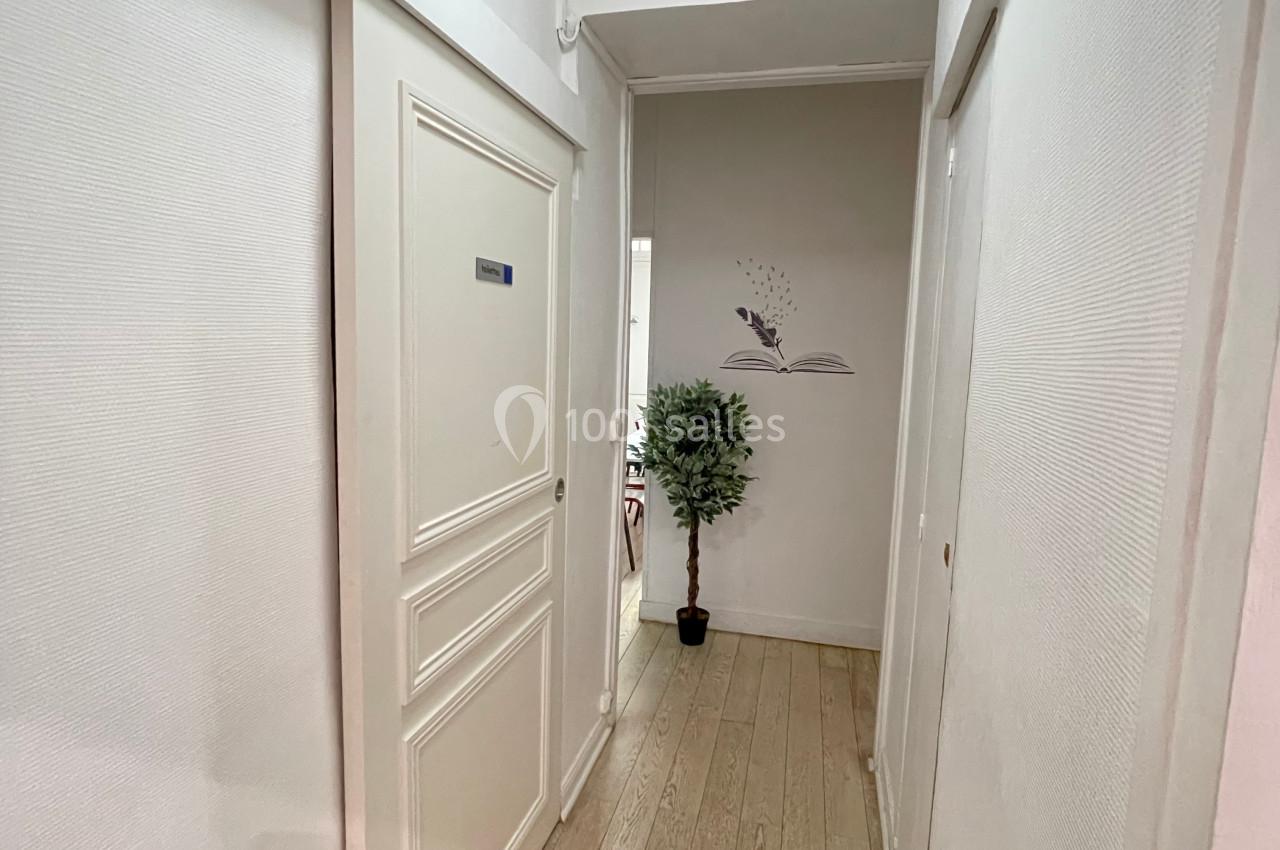Couloir étroit avec parquet clair, murs blancs, une porte à gauche et une plante en pot près d'un mur décoré.
