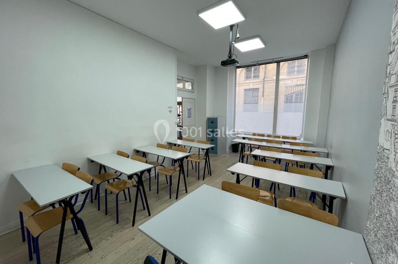 Salle de classe lumineuse avec des tables et chaises alignées, éclairée par des plafonniers et une grande fenêtre.