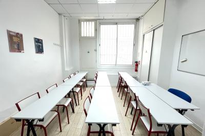 Salle de classe lumineuse avec des tables et chaises noires alignées, un projecteur suspendu et une grande fenêtre.