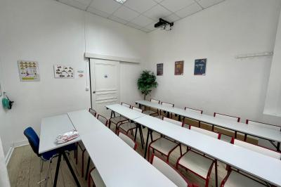 Salle de classe lumineuse avec des tables et chaises noires alignées, un projecteur suspendu et une grande fenêtre.