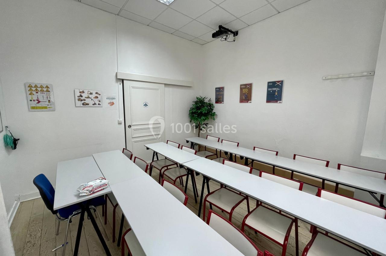 Salle de classe avec tables blanches alignées, chaises rouges, affiches murales et un projecteur au plafond.