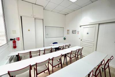 Salle de classe lumineuse avec des tables et chaises noires alignées, un projecteur suspendu et une grande fenêtre.
