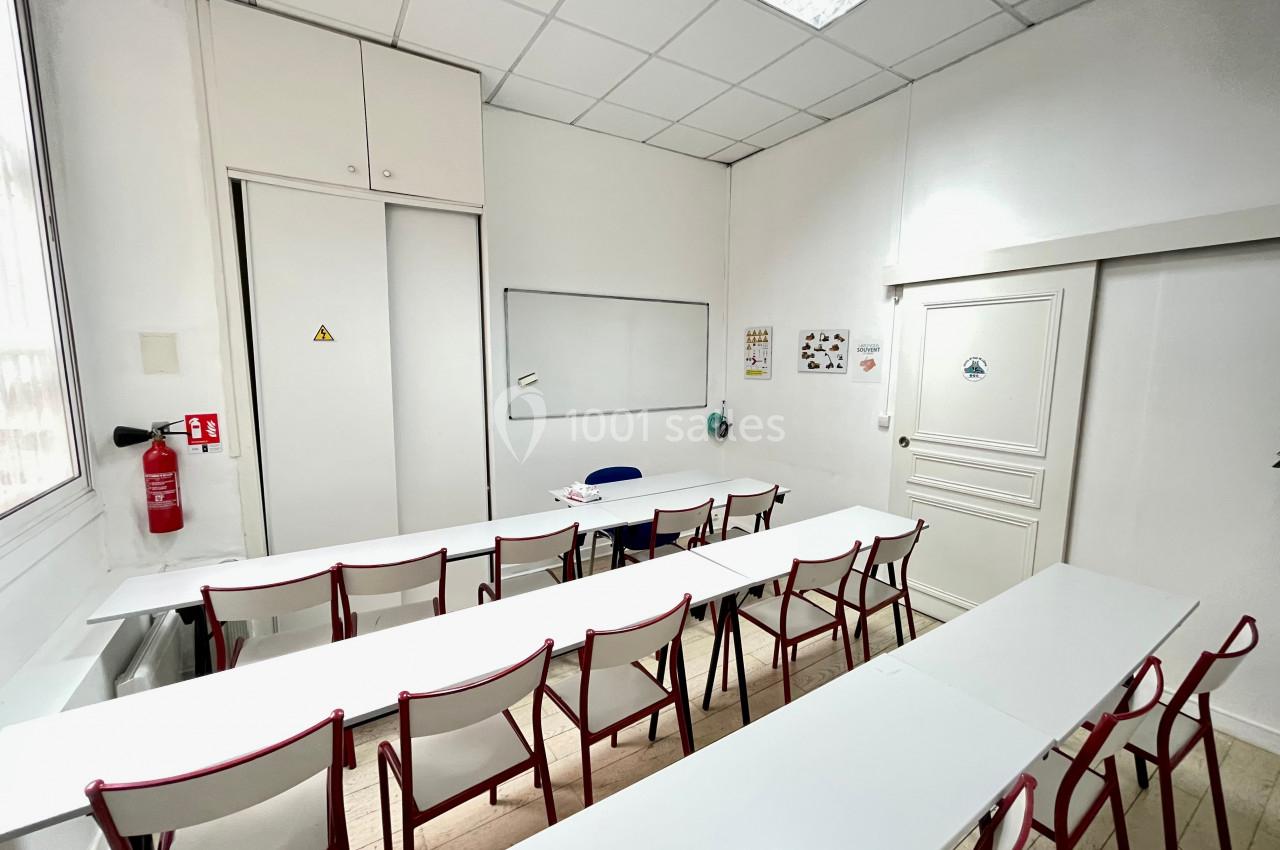 Salle de classe vide avec des tables blanches, des chaises rouges, un tableau blanc et une porte fermée.