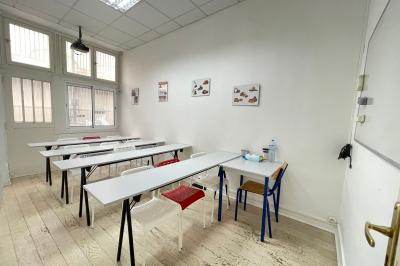 Salle de classe lumineuse avec des tables et chaises noires alignées, un projecteur suspendu et une grande fenêtre.
