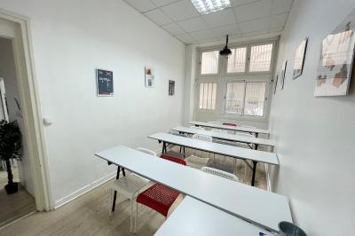 Salle de classe lumineuse avec des tables et chaises noires alignées, un projecteur suspendu et une grande fenêtre.