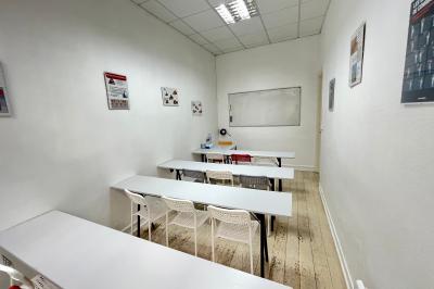Salle de classe lumineuse avec des tables et chaises noires alignées, un projecteur suspendu et une grande fenêtre.
