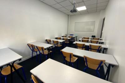 Salle de classe lumineuse avec des tables et chaises noires alignées, un projecteur suspendu et une grande fenêtre.
