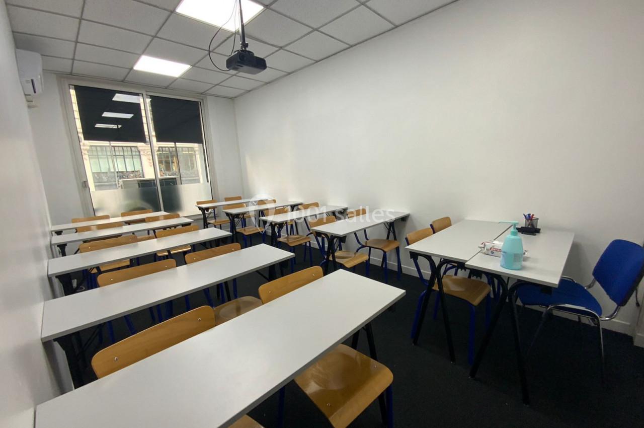 Salle de classe lumineuse avec des tables blanches, des chaises en bois et un projecteur suspendu au plafond.