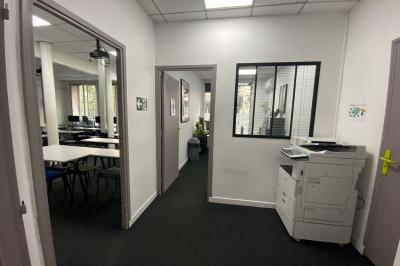 Salle de classe lumineuse avec des tables et chaises noires alignées, un projecteur suspendu et une grande fenêtre.