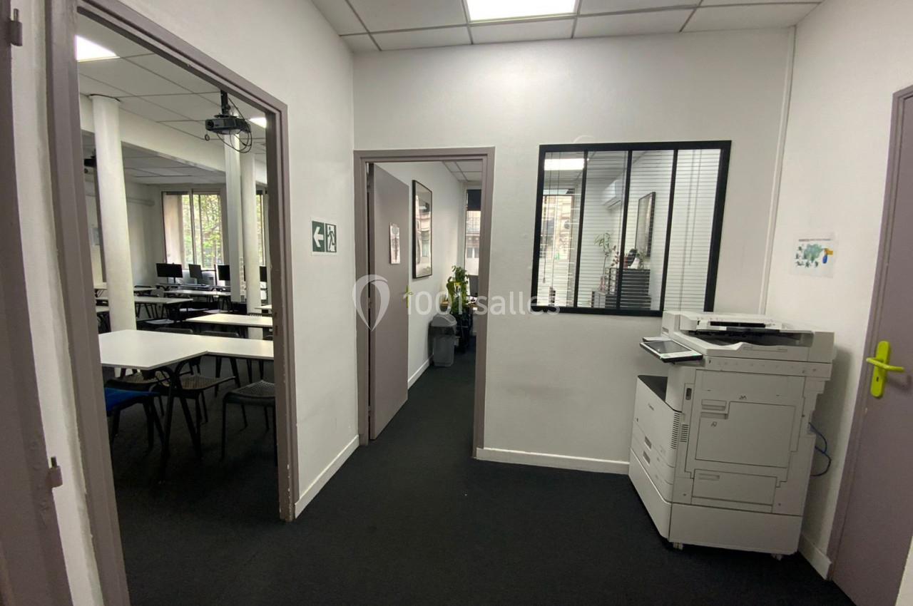 Couloir d'un espace de bureaux avec une imprimante à droite et une salle de réunion visible à gauche.