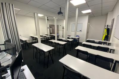 Salle de classe lumineuse avec des tables et chaises noires alignées, un projecteur suspendu et une grande fenêtre.