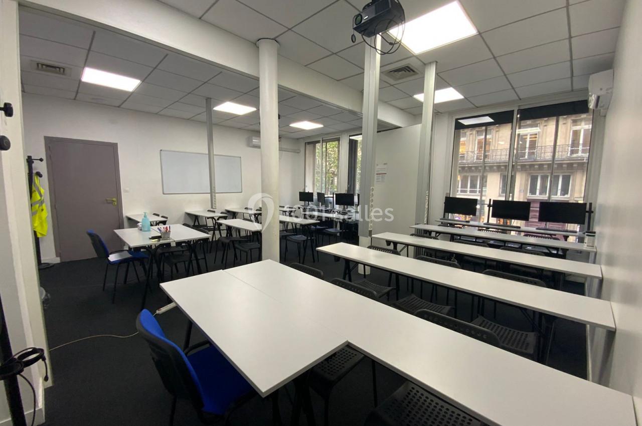 Salle de formation lumineuse avec des tables blanches, des chaises bleues et noires, et des ordinateurs alignés près des…