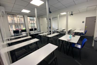 Salle de classe lumineuse avec des tables et chaises noires alignées, un projecteur suspendu et une grande fenêtre.