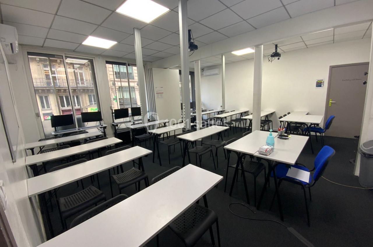 Salle de formation lumineuse avec des tables blanches, des chaises noires et des ordinateurs près des fenêtres.