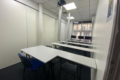 Salle de classe lumineuse avec des tables et chaises noires alignées, un projecteur suspendu et une grande fenêtre.
