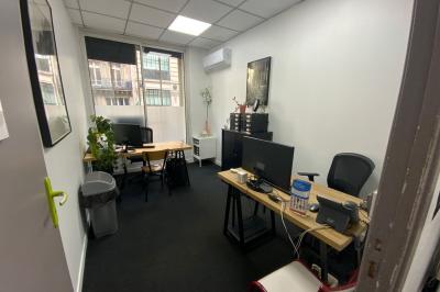 Salle de classe lumineuse avec des tables et chaises noires alignées, un projecteur suspendu et une grande fenêtre.