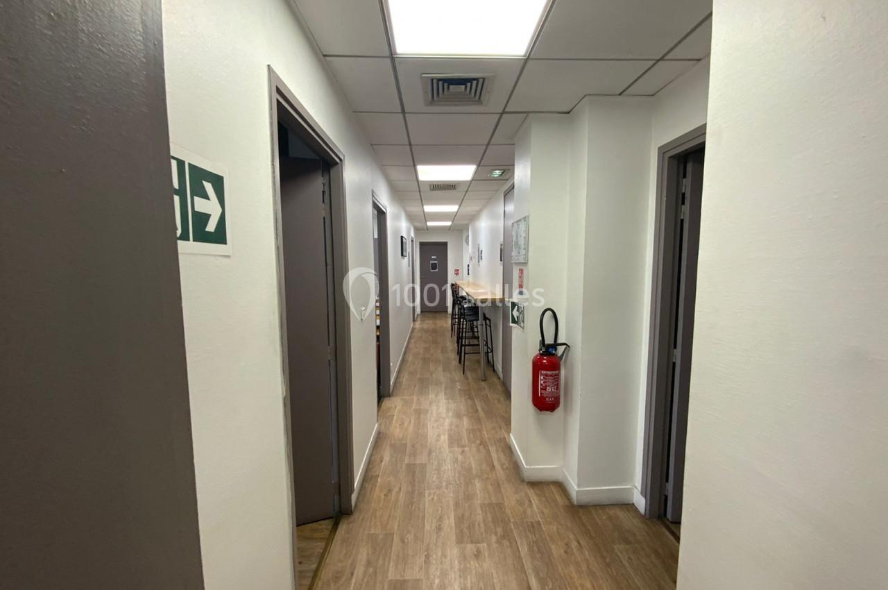 Couloir lumineux avec sol en bois, portes fermées, extincteur mural et signalisation de sortie sur la gauche.