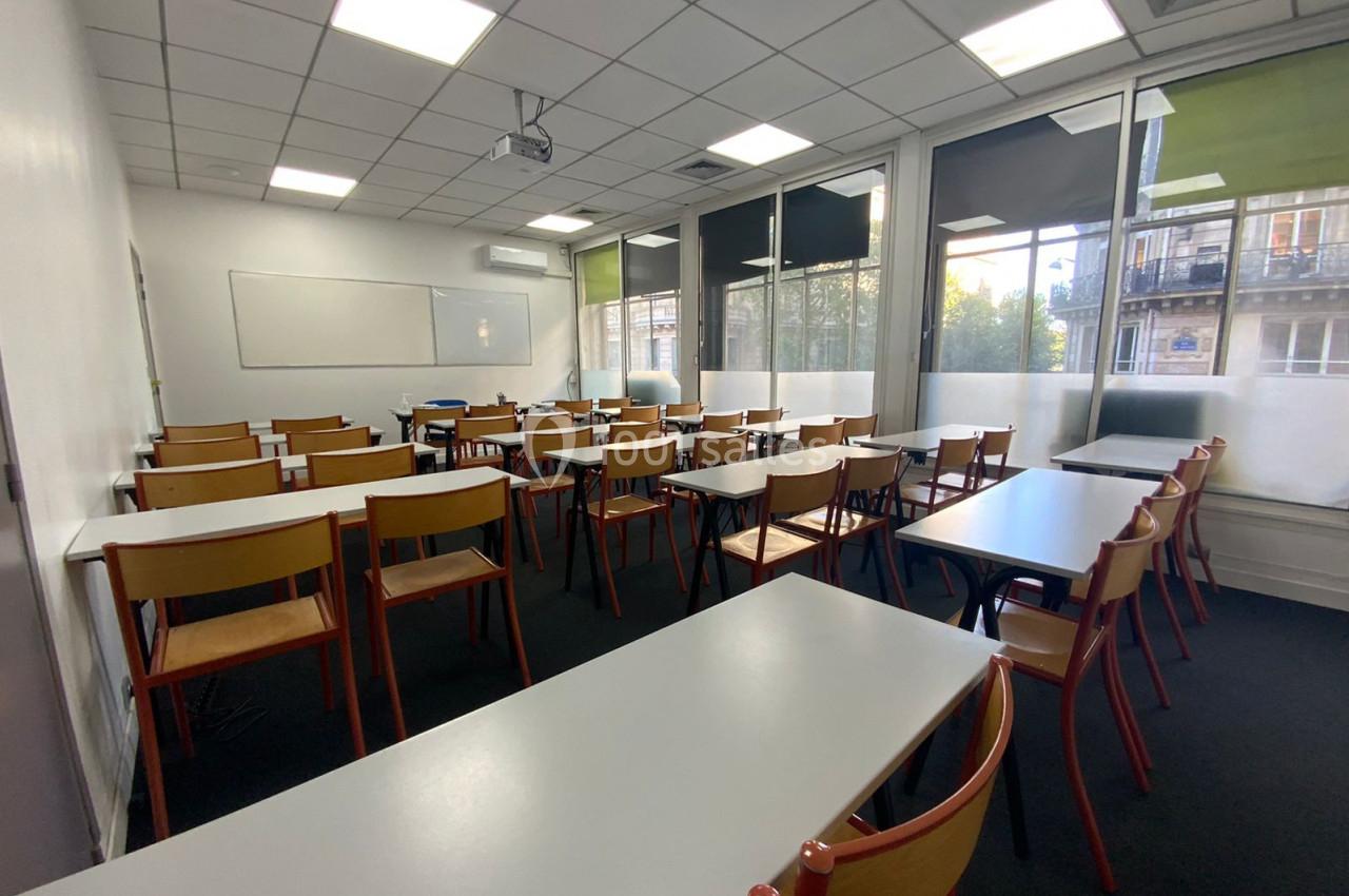Salle de classe vide avec des tables et chaises alignées, tableau blanc au mur et grandes fenêtres laissant entrer la…