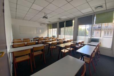 Salle de classe lumineuse avec des tables et chaises noires alignées, un projecteur suspendu et une grande fenêtre.