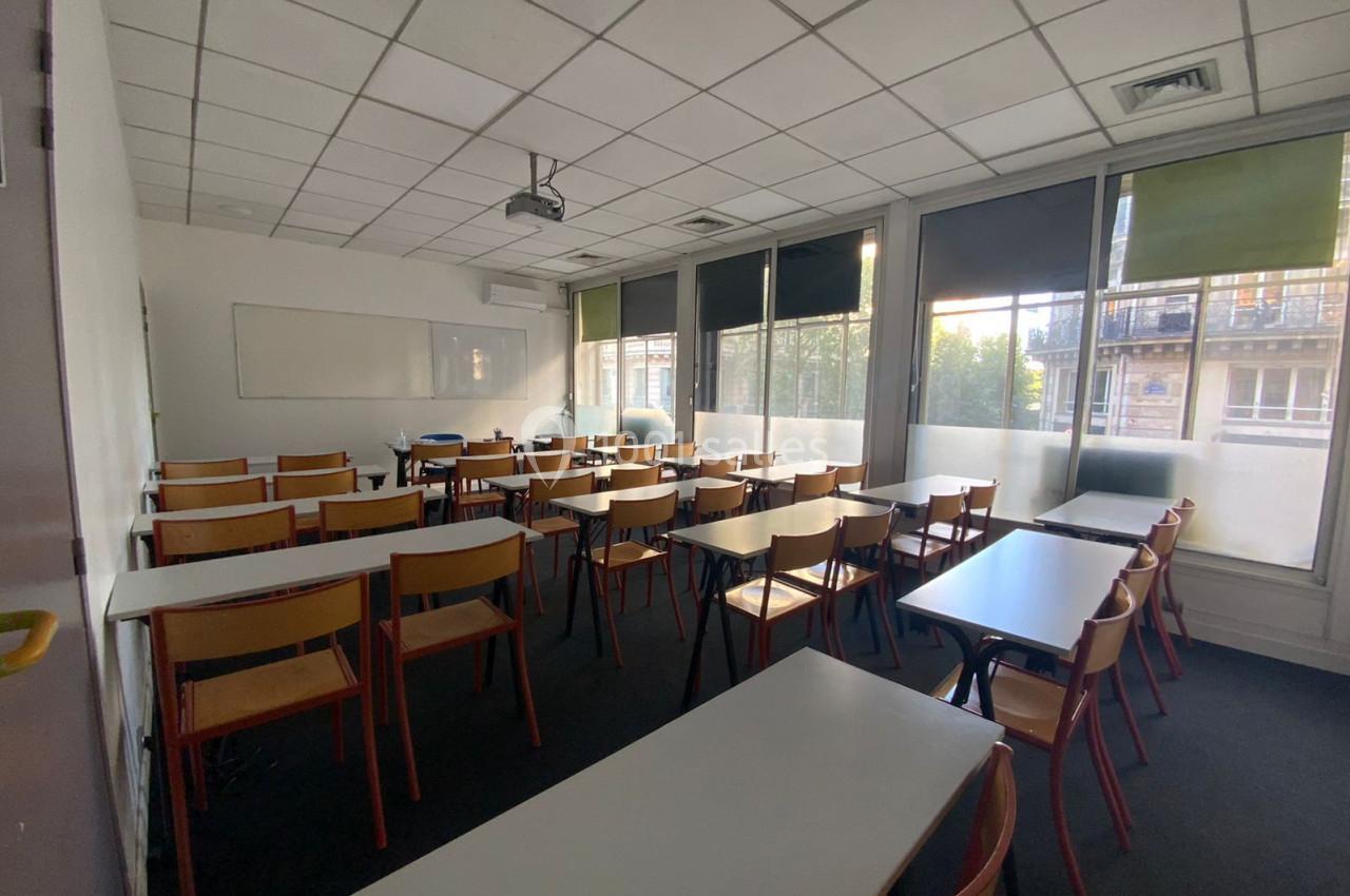 Salle de classe lumineuse avec des rangées de tables et chaises en bois, tableau blanc et projecteur au plafond.