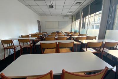 Salle de classe lumineuse avec des tables et chaises noires alignées, un projecteur suspendu et une grande fenêtre.