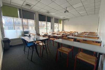 Salle de classe lumineuse avec des tables et chaises noires alignées, un projecteur suspendu et une grande fenêtre.