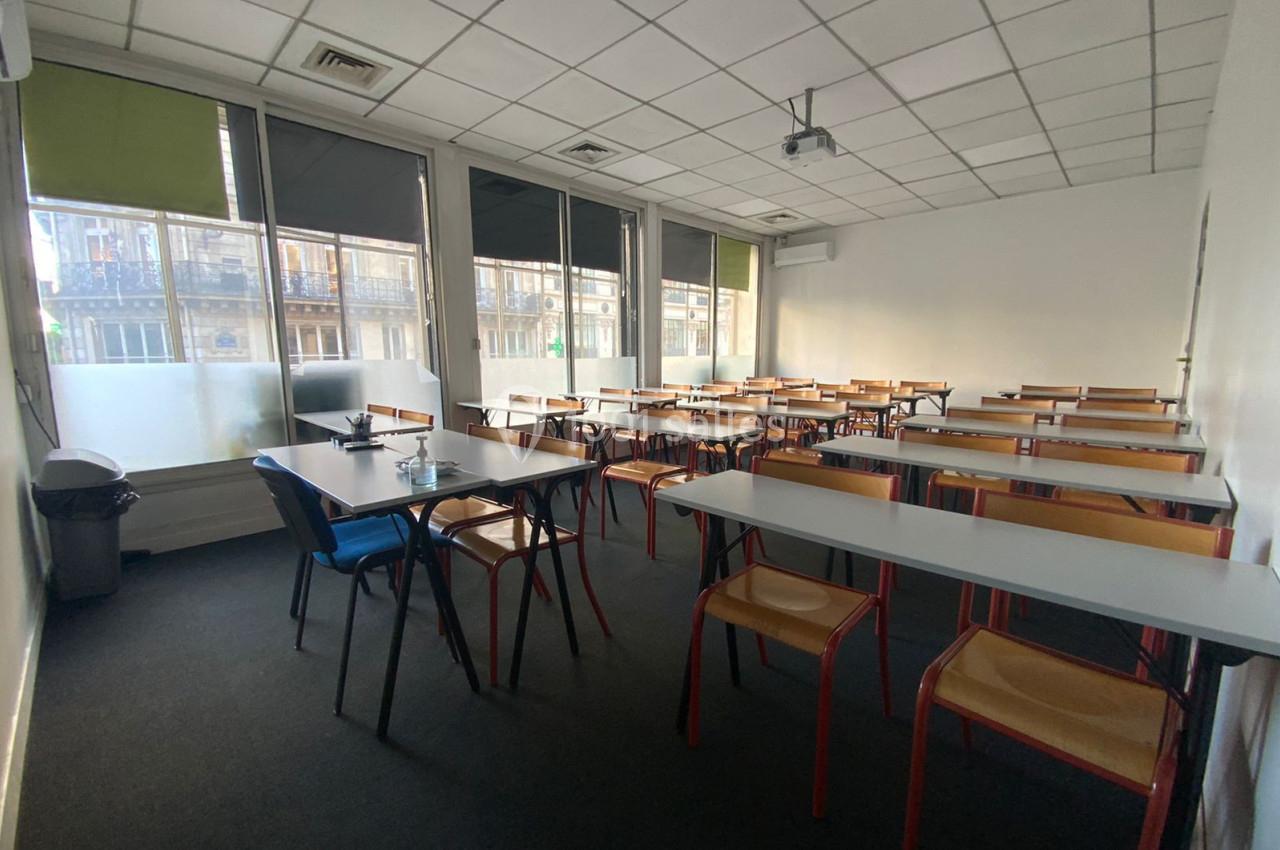 Salle de classe vide avec tables, chaises en bois, fenêtres donnant sur l'extérieur et plafond avec éclairage intégré.