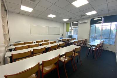 Salle de classe lumineuse avec des tables et chaises noires alignées, un projecteur suspendu et une grande fenêtre.