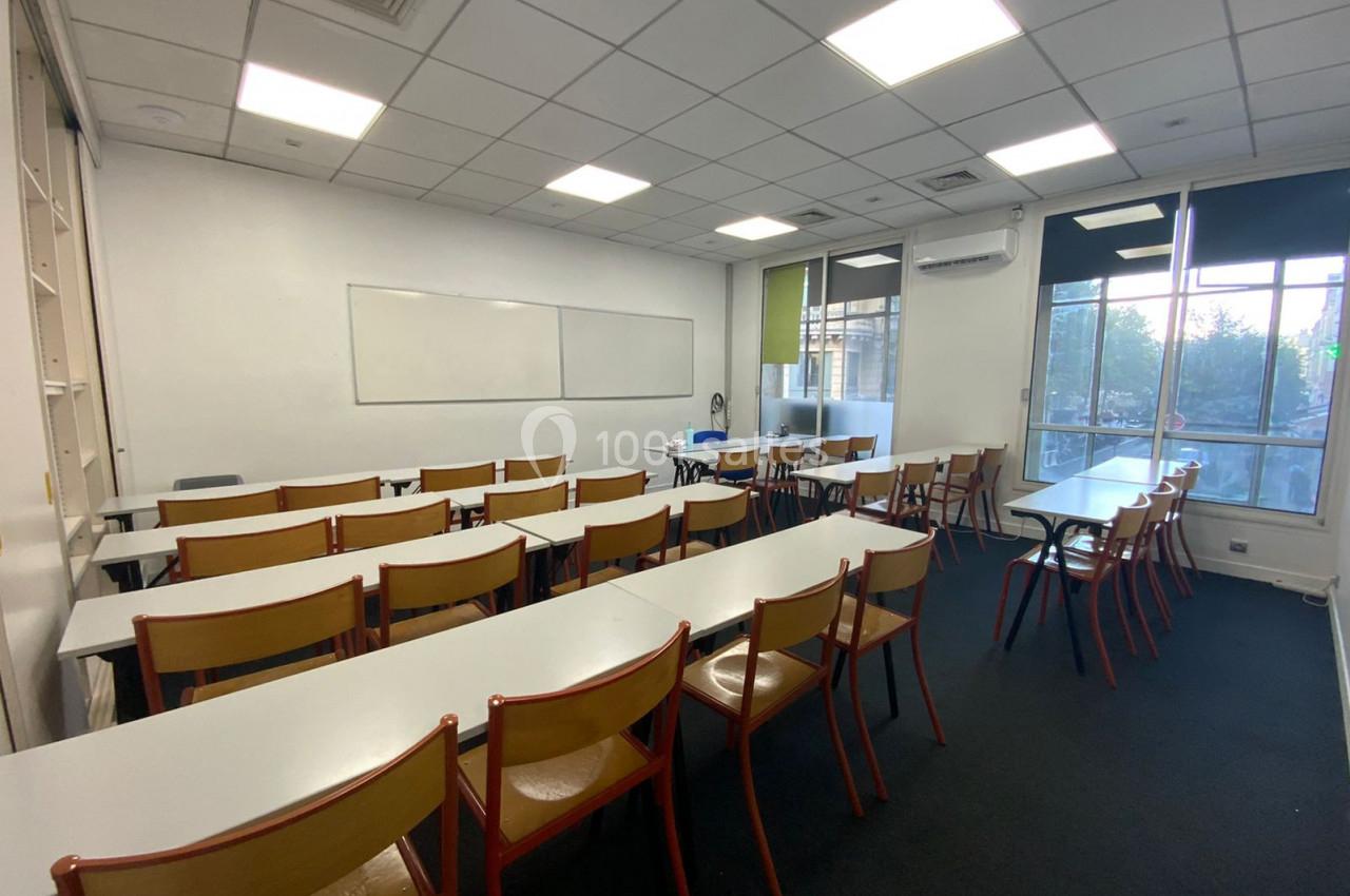 Salle de classe vide avec des rangées de tables et chaises, un tableau blanc et des fenêtres laissant entrer la lumière…