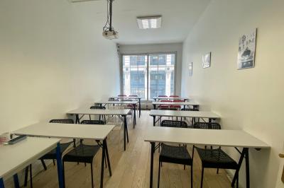 Salle de classe lumineuse avec des tables et chaises noires alignées, un projecteur suspendu et une grande fenêtre.
