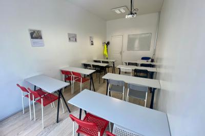 Salle de classe lumineuse avec des tables et chaises noires alignées, un projecteur suspendu et une grande fenêtre.
