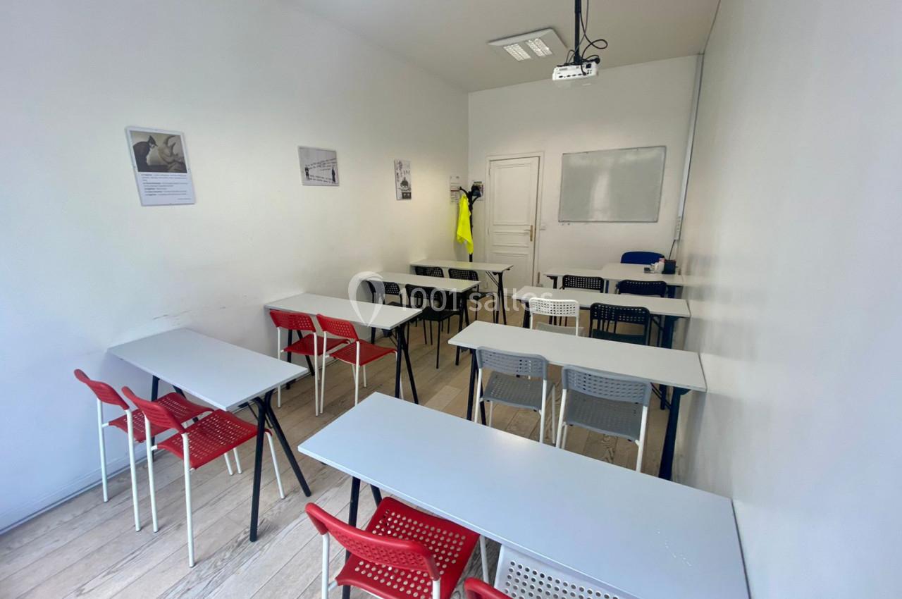 Salle de formation avec des tables blanches, des chaises rouges et noires, un tableau blanc et un projecteur au plafond.