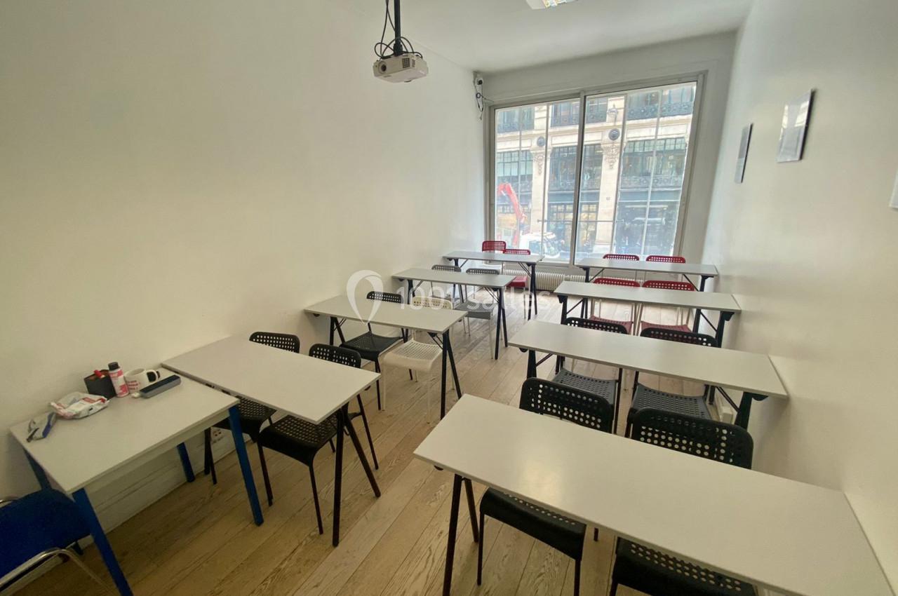 Salle de classe lumineuse avec des tables blanches, des chaises noires et une grande fenêtre donnant sur la rue.