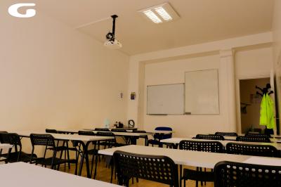 Salle de classe lumineuse avec des tables et chaises noires alignées, un projecteur suspendu et une grande fenêtre.