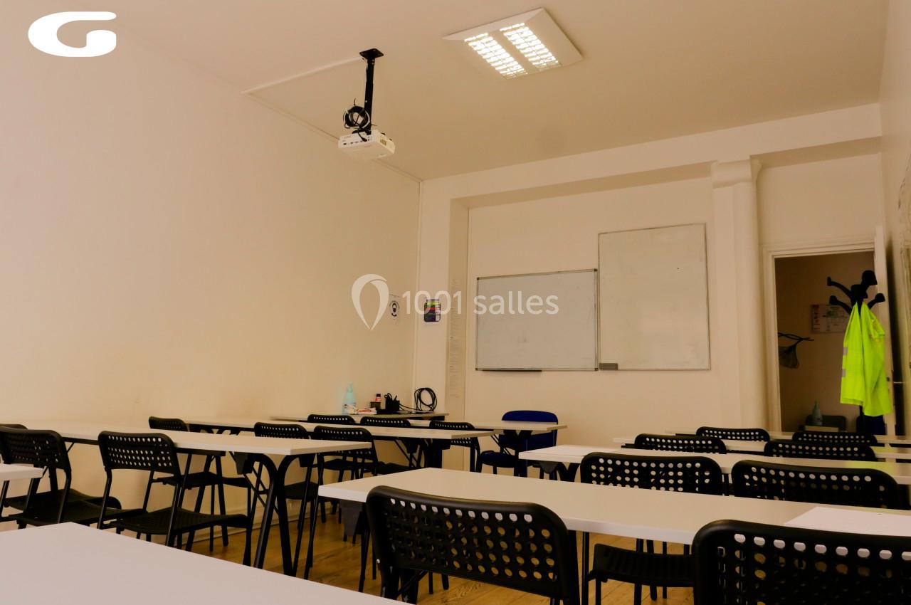 Salle de classe vide avec des tables, des chaises noires, un tableau blanc et un projecteur au plafond.