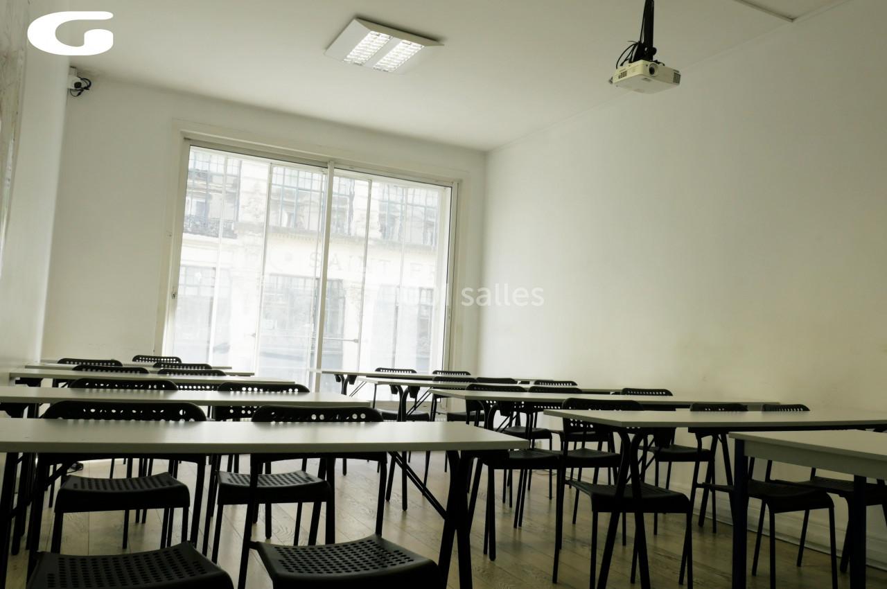 Salle de classe lumineuse avec des tables et chaises noires alignées, un projecteur suspendu et une grande fenêtre.