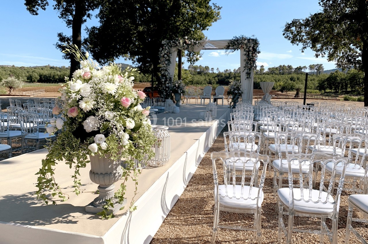 Allée décorée de fleurs blanches et roses menant à une arche de mariage sous des arbres, avec des chaises alignées.