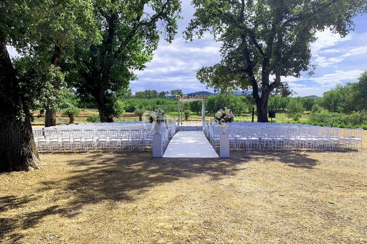 Allée centrale bordée de chaises blanches menant à une arche décorée de fleurs, dans un cadre extérieur arboré.