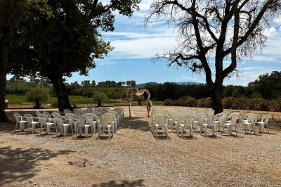 Allée centrale bordée de chaises blanches menant à une arche décorée de fleurs, dans un cadre extérieur arboré.