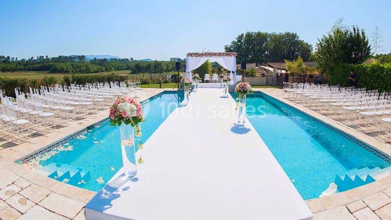 Allée blanche décorée pour une cérémonie en plein air, bordée d'une piscine et de chaises transparentes.
