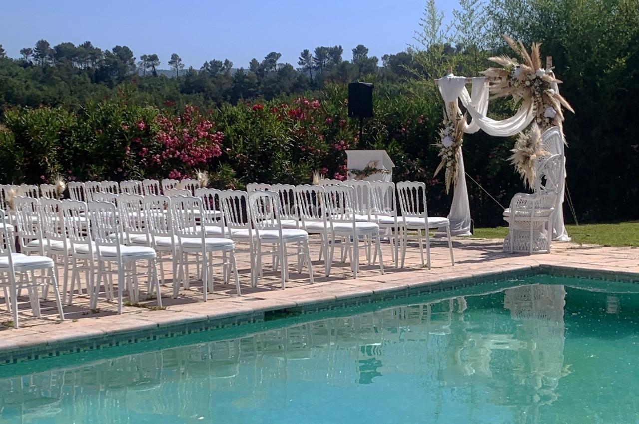 Chaises blanches alignées près d'une piscine, face à une arche décorée pour une cérémonie en extérieur.