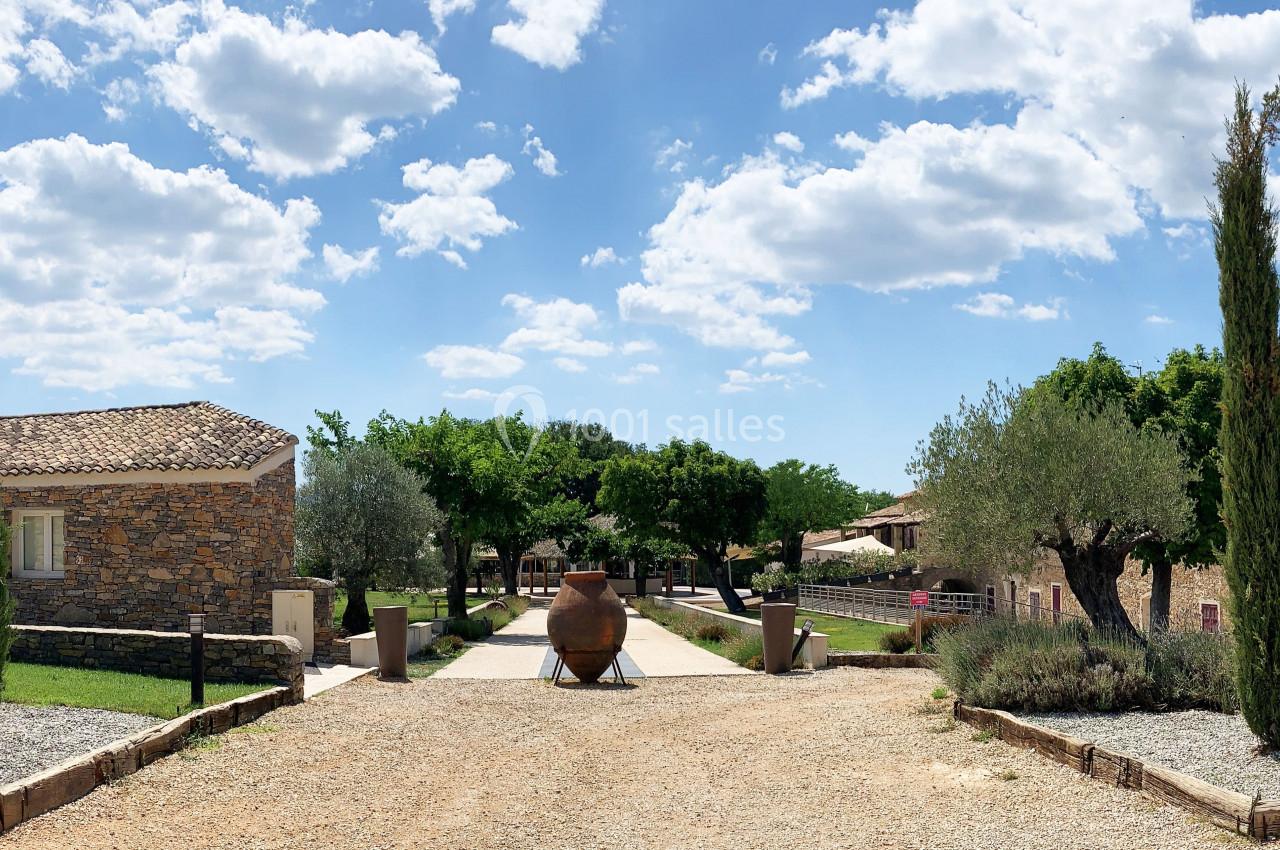 Vue d'une allée bordée d'arbres et de bâtiments en pierre, avec des amphores décoratives et un ciel dégagé.