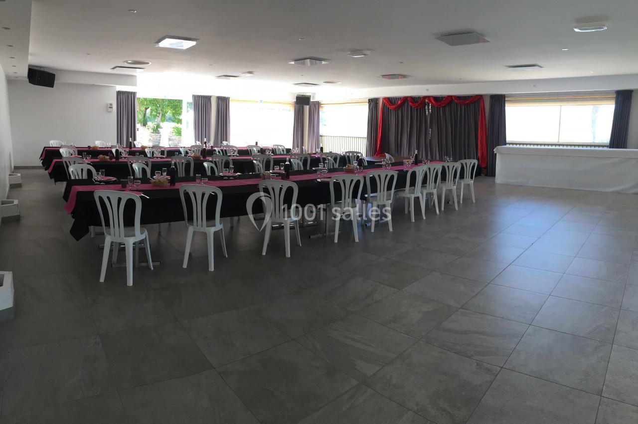 Salle de réception lumineuse avec tables dressées en noir et rose, chaises blanches et rideaux violets.