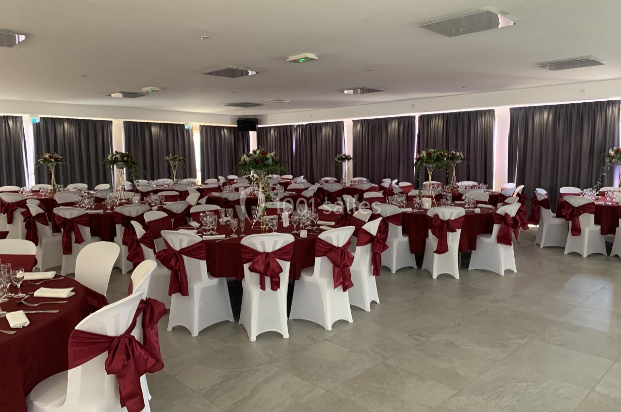 Salle de réception décorée avec des tables rondes, nappes rouges, chaises blanches et centres de table fleuris.
