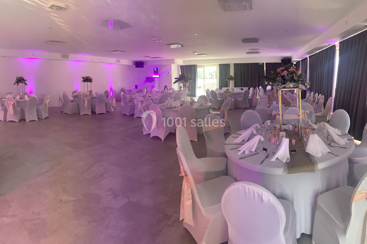 Salle de réception décorée avec des tables rondes, nappes blanches, chaises houssées et éclairage d'ambiance rose.
