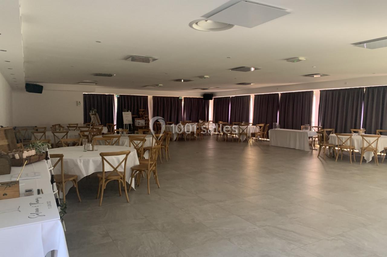 Salle spacieuse avec tables rondes et chaises en bois, éclairée par des fenêtres avec rideaux sombres.