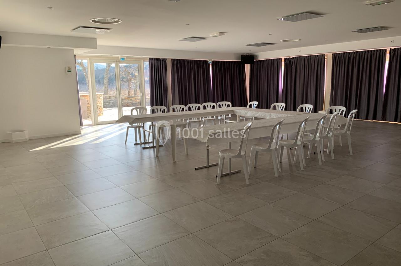 Salle lumineuse avec des tables disposées en U, entourées de chaises blanches, et des rideaux fermés sur les fenêtres.