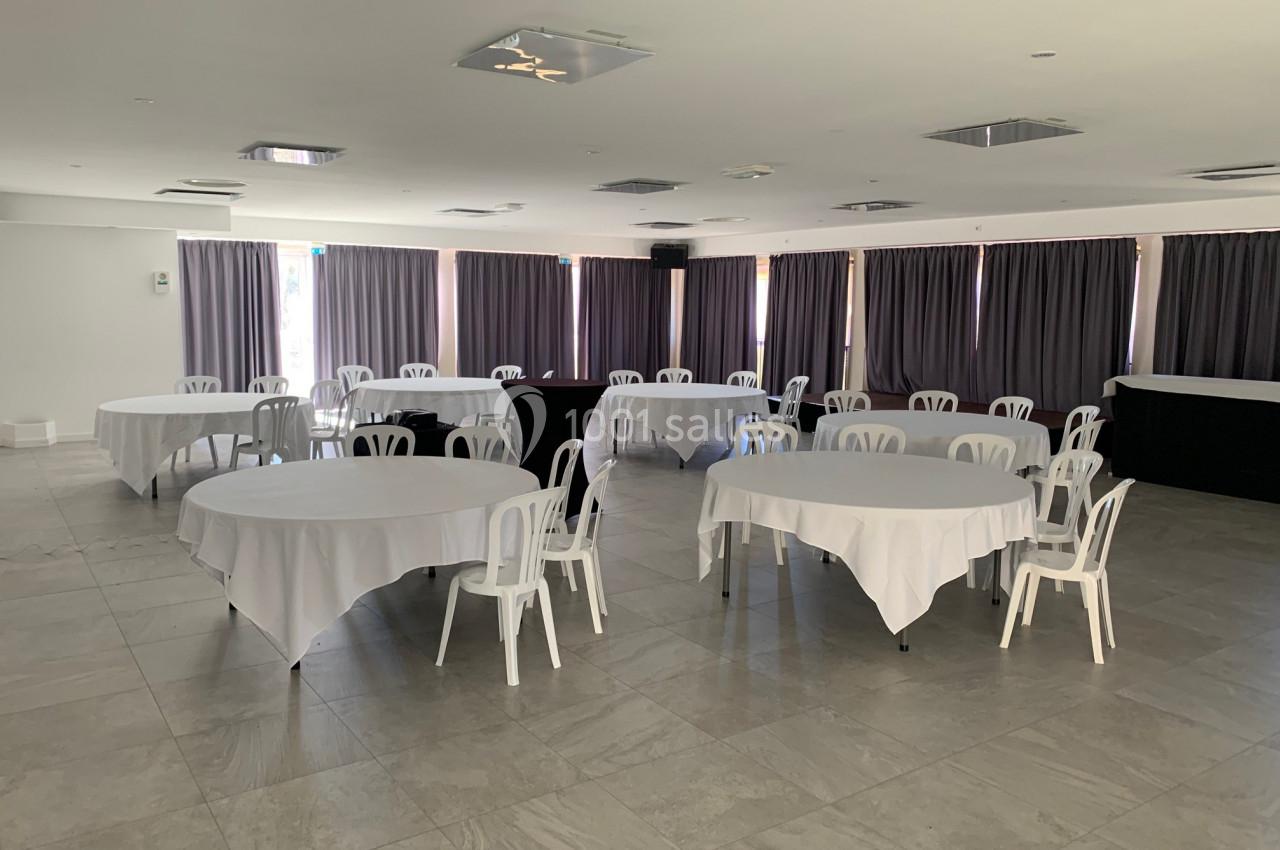 Salle de réception lumineuse avec tables rondes recouvertes de nappes blanches et chaises en plastique blanc.