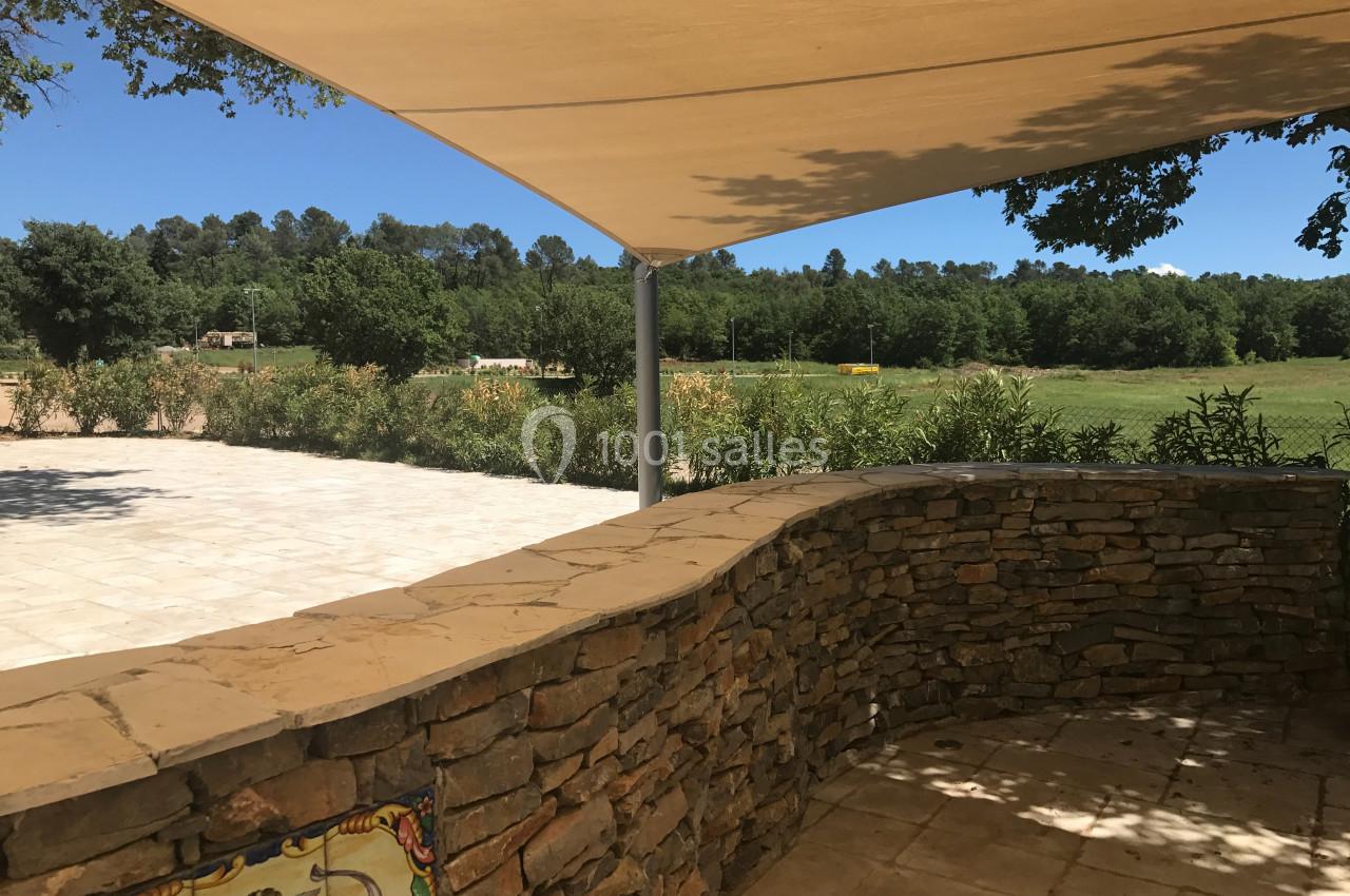 Mur en pierre courbé sous une toile d'ombrage, avec vue sur un espace vert et des arbres en arrière-plan.