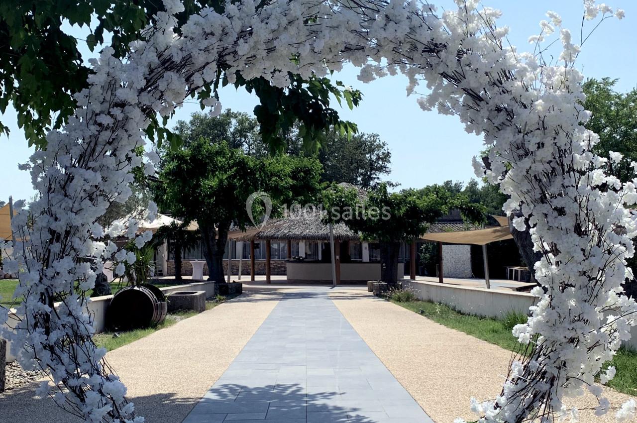 Arche décorée de fleurs blanches sur une allée pavée, entourée d'arbres et de bâtiments en arrière-plan.